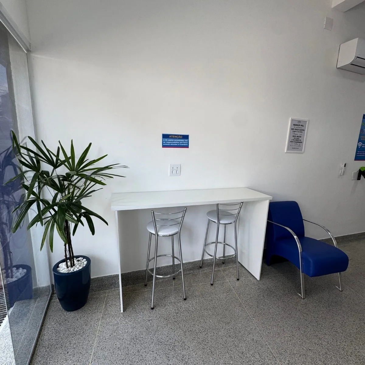 Área de descanso da Tá Limpo! Lavanderia com bancada branca, banquetas e planta decorativa