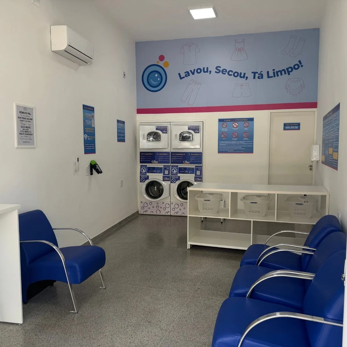 Interior da Tá Limpo! Lavanderia com poltronas azuis, ar-condicionado e painel Lavou, Secou, Tá Limpo